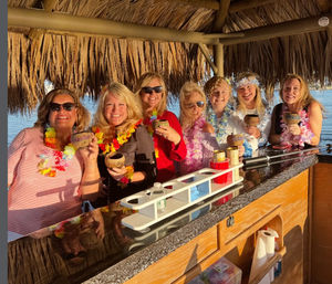 Seven friends in leis smiling at a waterfront tiki bar, holding decorated coconut cups and tropical drinks under a thatched palapa at sunset.