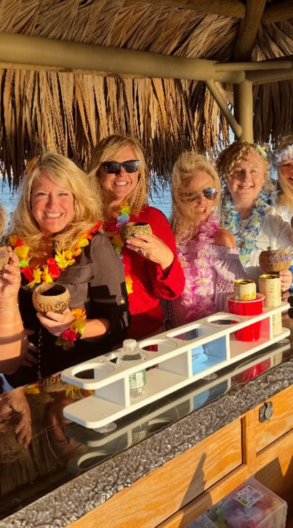 Seven friends in leis smiling at a waterfront tiki bar, holding decorated coconut cups and tropical drinks under a thatched palapa at sunset.
