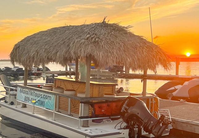 Tiki-style pontoon boat with thatched palapa roof docked at a coastal marina during a golden sunset, jet skis and outboard motor visible on calm water.