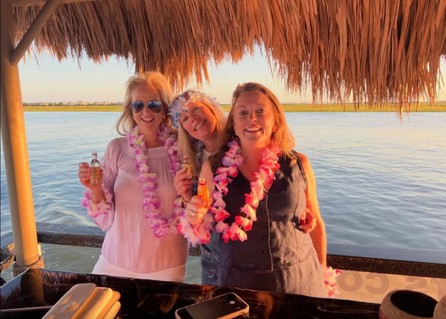 Three smiling women wearing pink and purple flower leis and holding small bottles under a tiki-style thatched roof, waterfront sunset over calm coastal marsh