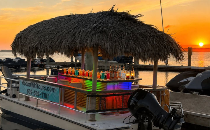 Tiki-style pontoon bar with a thatched roof and rainbow-lit counter lined with colorful cocktails at a marina sunset over calm water