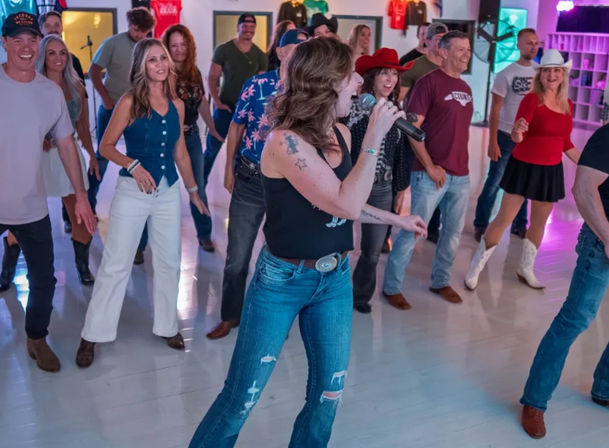 Country line dance class in a bright studio: adults in cowboy hats, boots and jeans following two microphone-toting instructors leading upbeat steps.