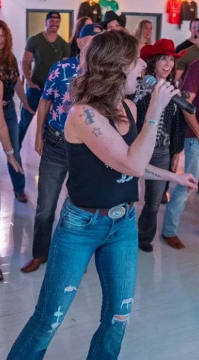 Country line dance class in a bright studio: adults in cowboy hats, boots and jeans following two microphone-toting instructors leading upbeat steps.