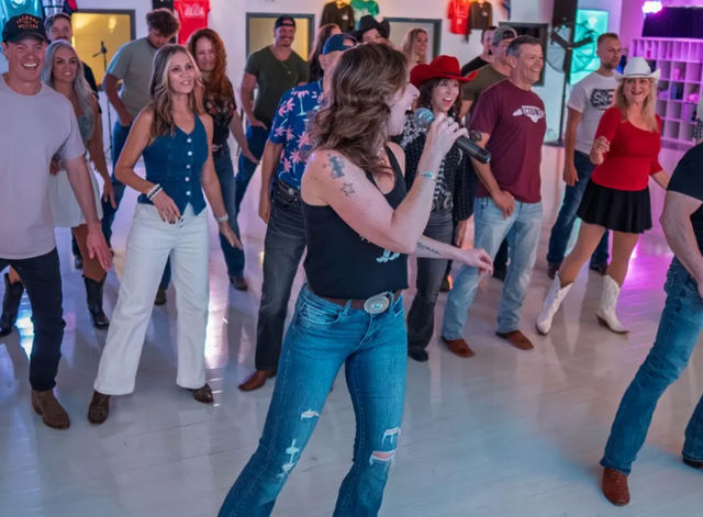 Country line dance class in a bright studio: adults in cowboy hats, boots and jeans following two microphone-toting instructors leading upbeat steps.