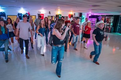 Lively country line dance in an indoor dance studio with a group wearing cowboy hats and boots, led by two singers holding microphones under colorful lights.