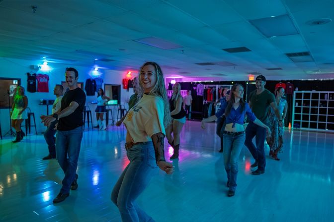 Smiling group of adults line-dancing in a neon-lit indoor studio with colorful blue and purple lights, casual jeans and boots.