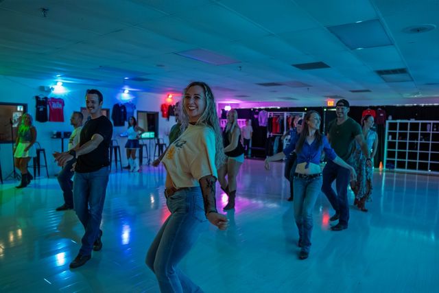 Smiling group of adults line-dancing in a neon-lit indoor studio with colorful blue and purple lights, casual jeans and boots.