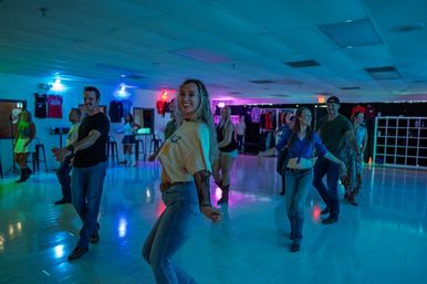 Smiling group doing a country-style line dance in a neon-lit indoor dance studio, wearing casual jeans and boots on a glossy floor
