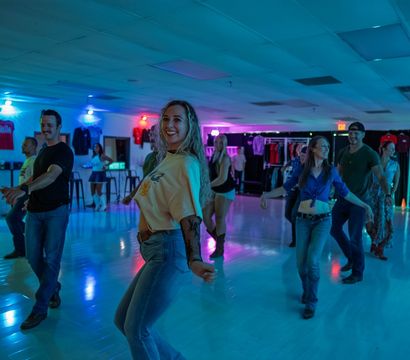 Smiling group doing a country-style line dance in a neon-lit indoor dance studio, wearing casual jeans and boots on a glossy floor