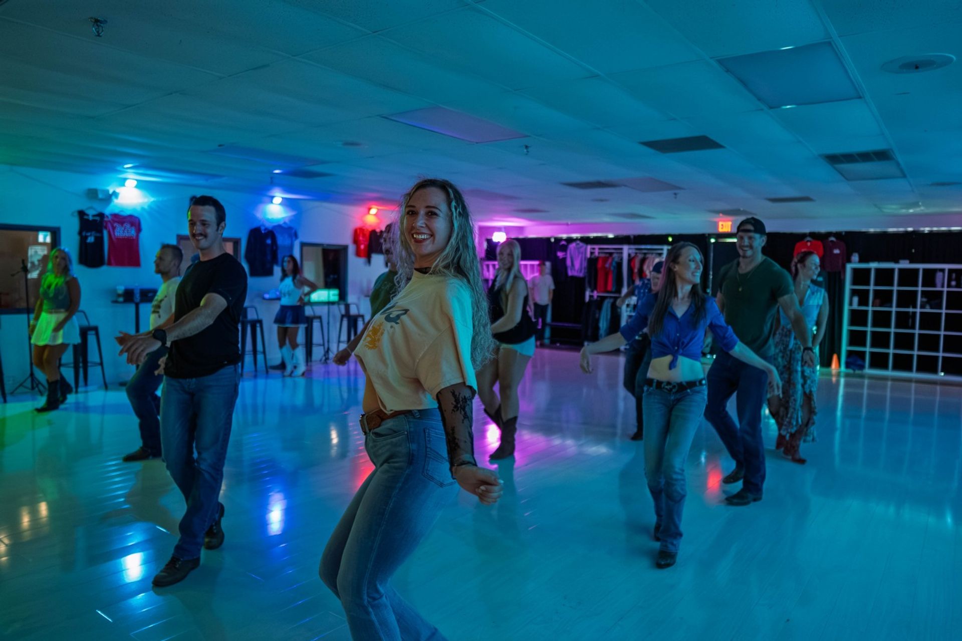 Smiling group doing a country-style line dance in a neon-lit indoor dance studio, wearing casual jeans and boots on a glossy floor