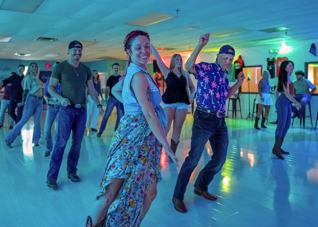 Adults enjoying a country line-dance class in a neon-lit indoor dance studio — woman in a floral skirt and man in a patterned shirt lead the group.