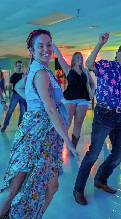 Adults enjoying a country line-dance class in a neon-lit indoor dance studio — woman in a floral skirt and man in a patterned shirt lead the group.