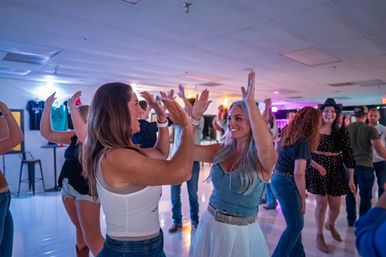 Two smiling women laugh and high-five on a crowded indoor dance floor as other party-goers dance under colorful lights — lively indoor nightlife scene.