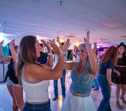 Two smiling women laugh and high-five on a crowded indoor dance floor as other party-goers dance under colorful lights — lively indoor nightlife scene.