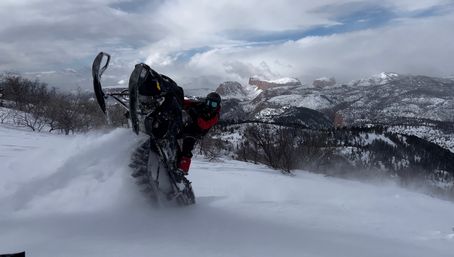Snowmobile kicking up powder as a rider does a wheelie on a snowy mountain slope with rugged snow-covered peaks and cliffs in the background.