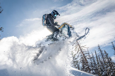 Snowmobile rider catching big air through deep powder in alpine backcountry, snow spraying beneath a bright blue sky and snow-covered pine trees.