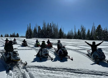 Sunlit snowfield with a group of snowmobiles and riders lined up, crisscrossing tracks in the snow, evergreen trees on the horizon and a clear blue sky; one rider raises their arms in celebration.