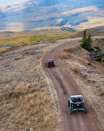 Two off-road UTVs cruising a winding mountain dirt road through golden autumn hills and a scenic valley.