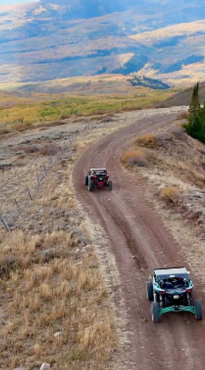Two off-road UTVs cruising a winding mountain dirt road through golden autumn hills and a scenic valley.