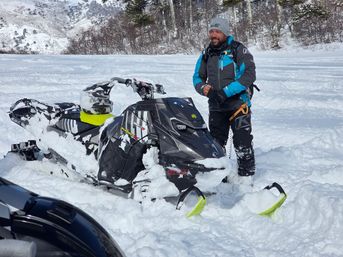 Smiling person in blue winter gear standing beside a black snowmobile with neon-yellow skis stuck in deep powder on a snowy mountain slope near pine trees