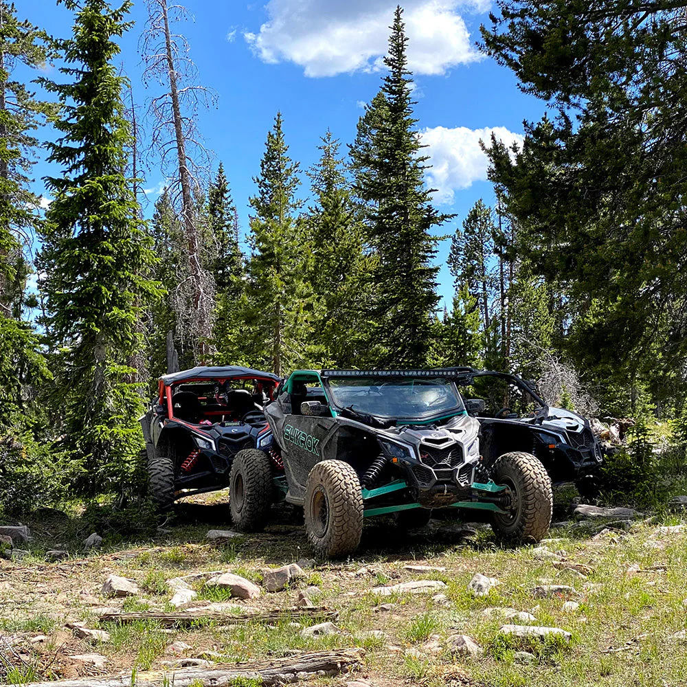 Two off-road UTV side-by-sides parked on a rocky mountain trail in a pine forest under a bright blue sky