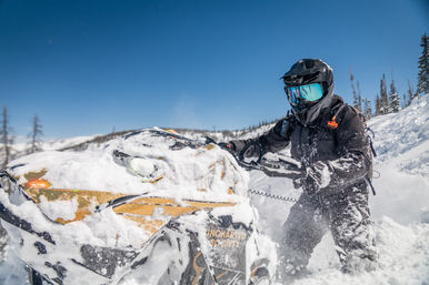 Snowmobile rider in black helmet and mirrored goggles powering through deep powder on a yellow snowmobile in sunny mountain backcountry under a clear blue sky