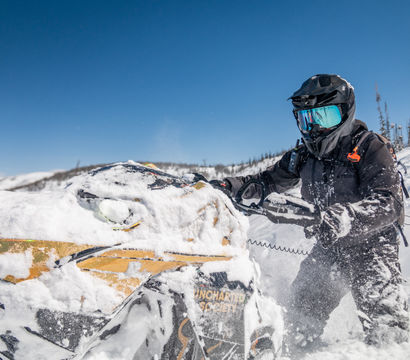 Snowmobile rider in black helmet and mirrored goggles powering through deep powder on a yellow snowmobile in sunny mountain backcountry under a clear blue sky