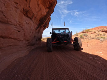 Rugged off-road UTV squeezing under a red sandstone overhang on a sunlit red‑rock desert trail, deep tire tracks in sandy soil and a bright blue sky overhead.