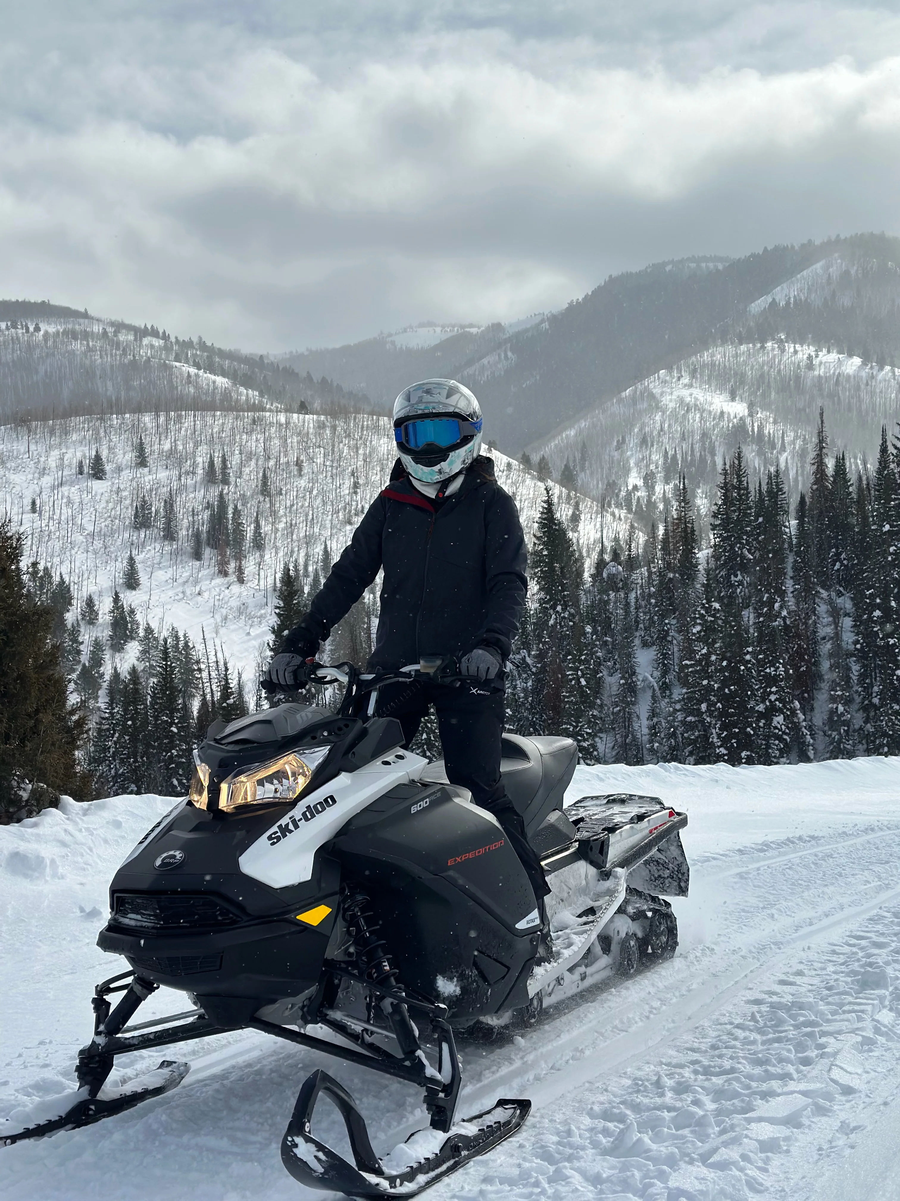 Rider in a blue-goggled helmet on a black-and-white snowmobile cruising a snowy alpine trail, pine-covered mountains and cloudy sky in the background.