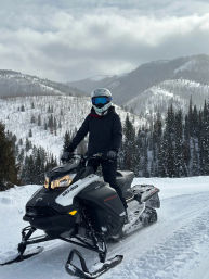 Rider in a blue-goggled helmet on a black-and-white snowmobile cruising a snowy alpine trail, pine-covered mountains and cloudy sky in the background.