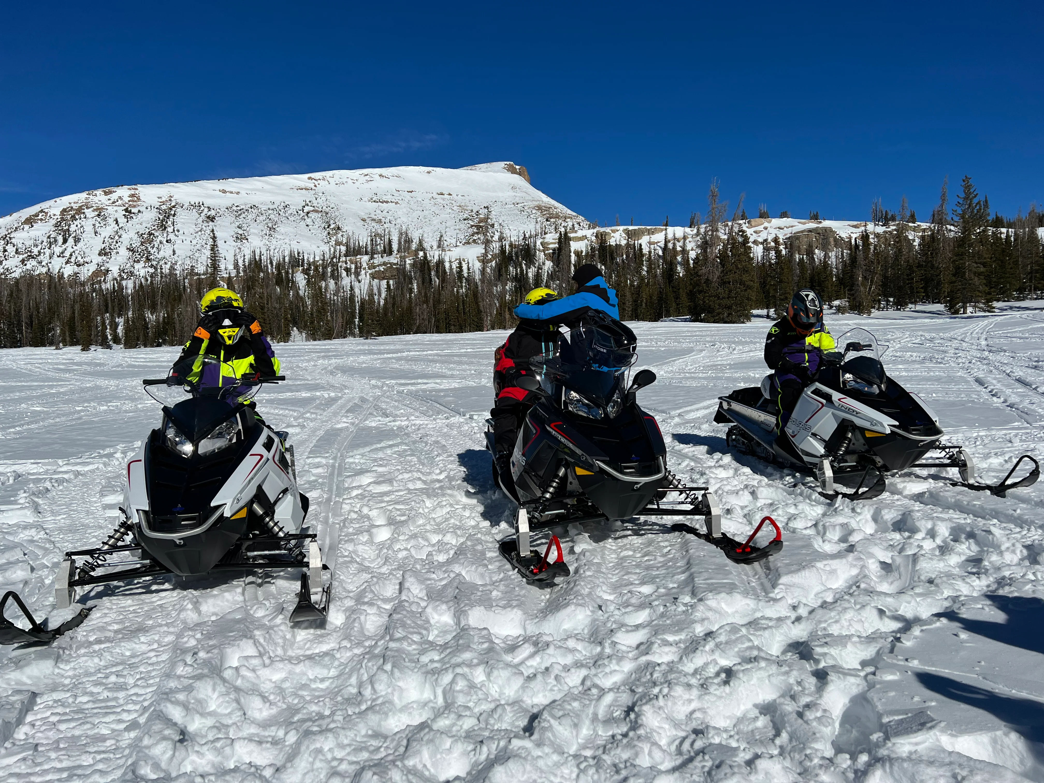 Three snowmobilers in bright jackets and helmets on parked snowmobiles across a sunlit alpine snowfield, with evergreen trees and a snow-covered mountain under a clear blue sky.