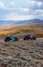 Two sporty off-road UTVs driving on a dirt trail through sagebrush with golden autumn hills and blue mountains under a dramatic cloudy sky