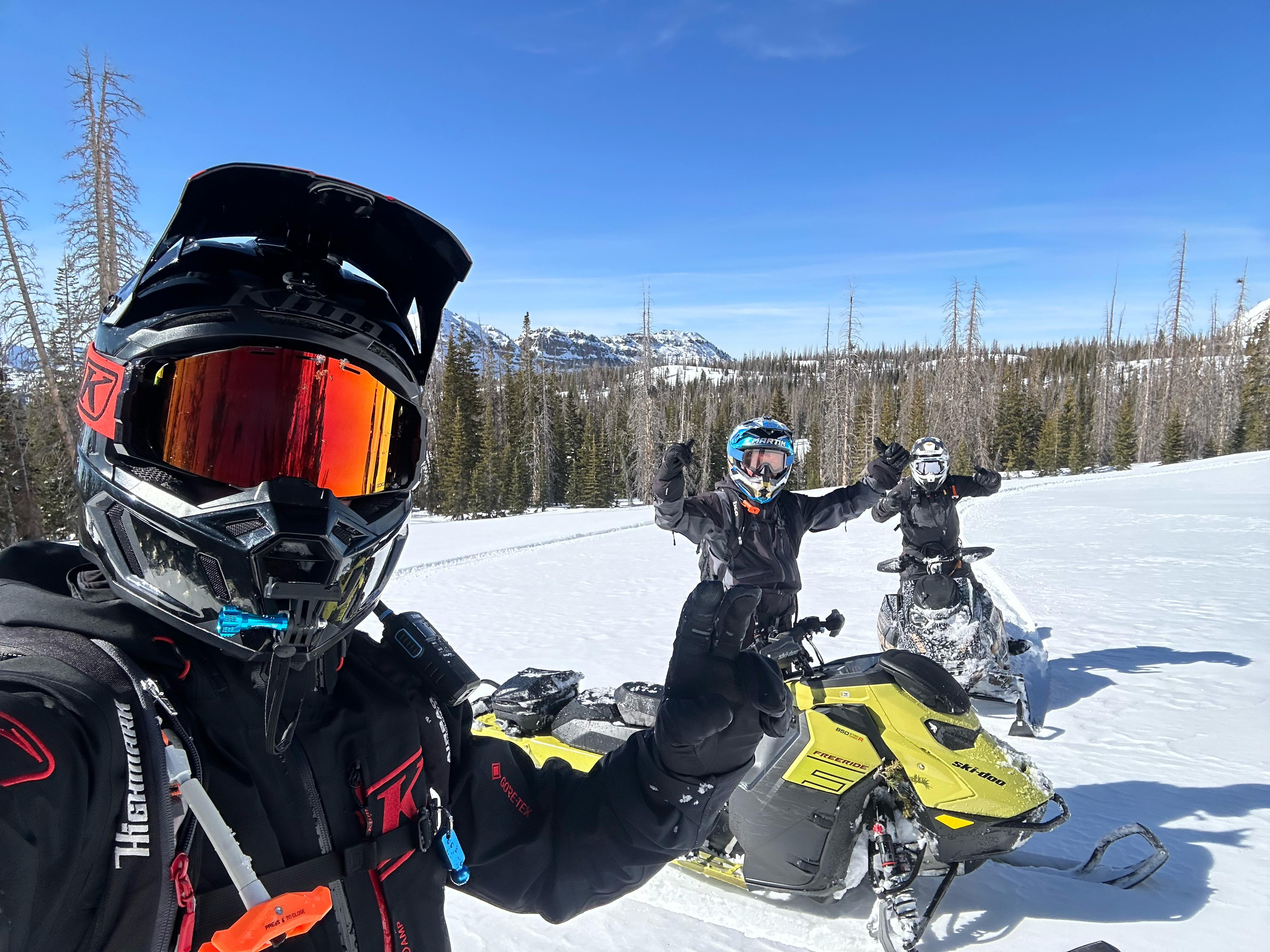 Three snowmobilers in helmets taking a selfie on a bright alpine snowfield, one on a yellow snowmobile with others cheering, surrounded by evergreen forest and distant snow-capped mountains under a clear blue sky