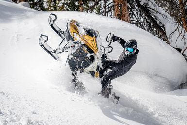 Snowmobiler in black gear and blue goggles lifts the front of a yellow snowmobile through deep powder near snow-covered trees in a mountain forest