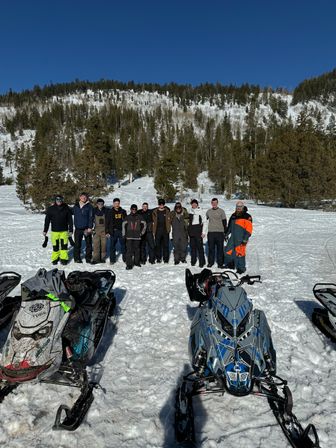 Group of snowmobilers posing behind parked sleds on a sunny snowfield — a winter snowmobiling adventure with pine‑covered mountain slopes and clear blue sky.