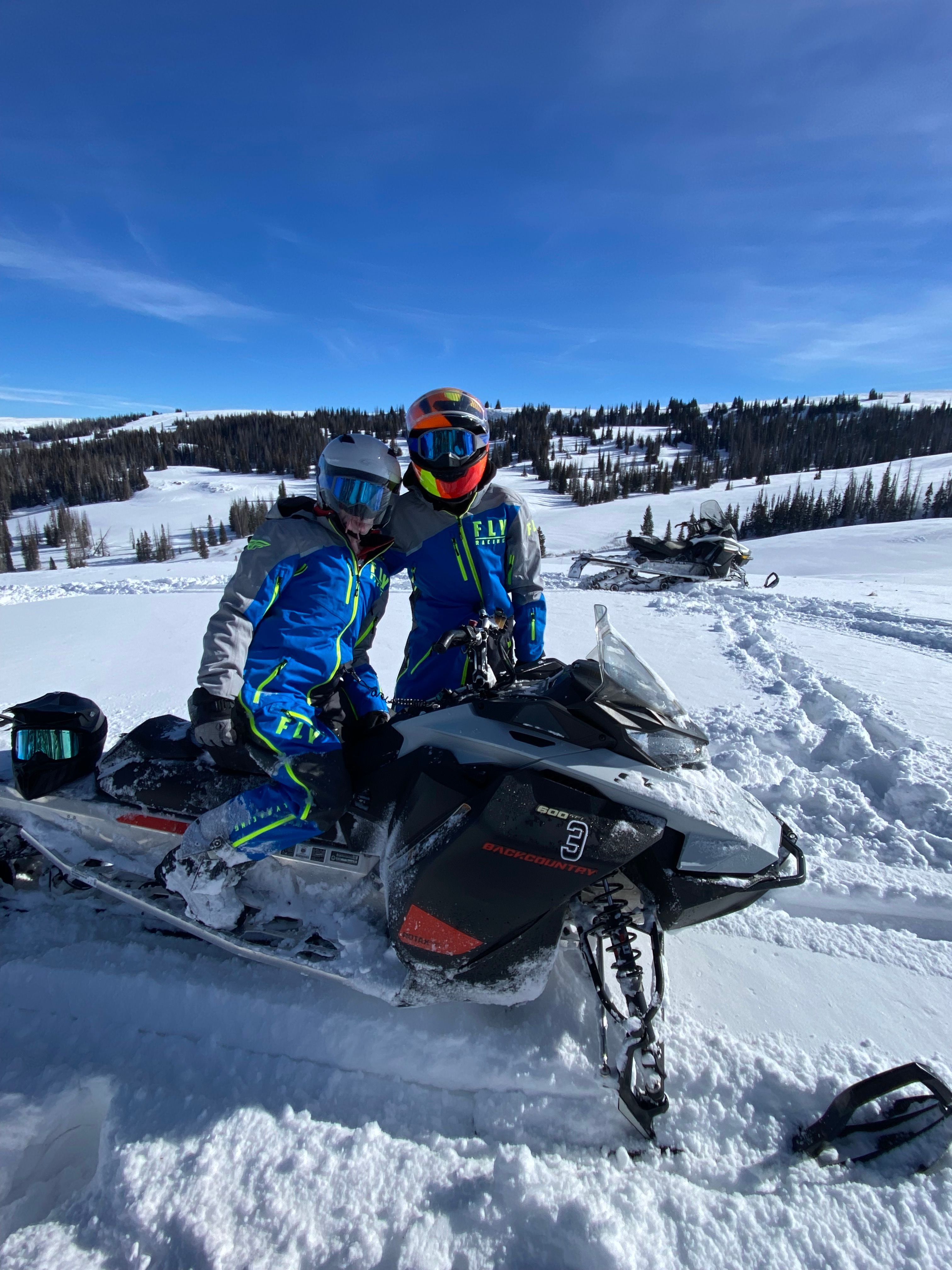 Two riders in bright blue snowmobile suits and reflective helmets posed on a black snowmobile in a snowy alpine meadow with pine trees and a clear blue sky — winter snowmobile scene.
