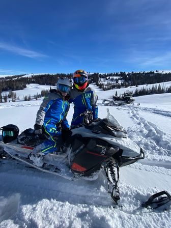 Two riders in bright blue snowmobile suits and reflective helmets posed on a black snowmobile in a snowy alpine meadow with pine trees and a clear blue sky — winter snowmobile scene.