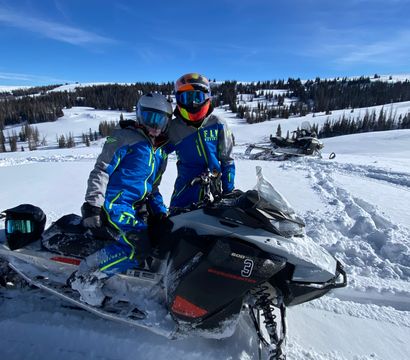 Two riders in bright blue snowmobile suits and reflective helmets posed on a black snowmobile in a snowy alpine meadow with pine trees and a clear blue sky — winter snowmobile scene.