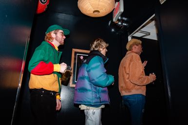 Three people in colorful jackets holding beer mugs, laughing as they enter a dim urban bar hallway beneath an illuminated EXIT sign.
