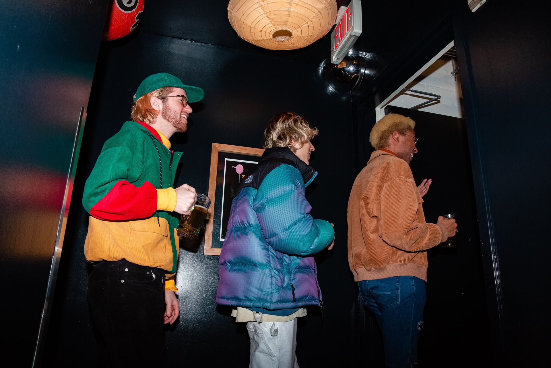 Three people in colorful jackets holding beer mugs, laughing as they enter a dim urban bar hallway beneath an illuminated EXIT sign.