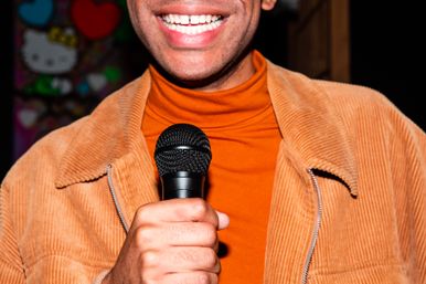 Close-up of a grinning performer holding a black microphone, wearing an orange turtleneck and tan corduroy jacket at an indoor open-mic/comedy setting.
