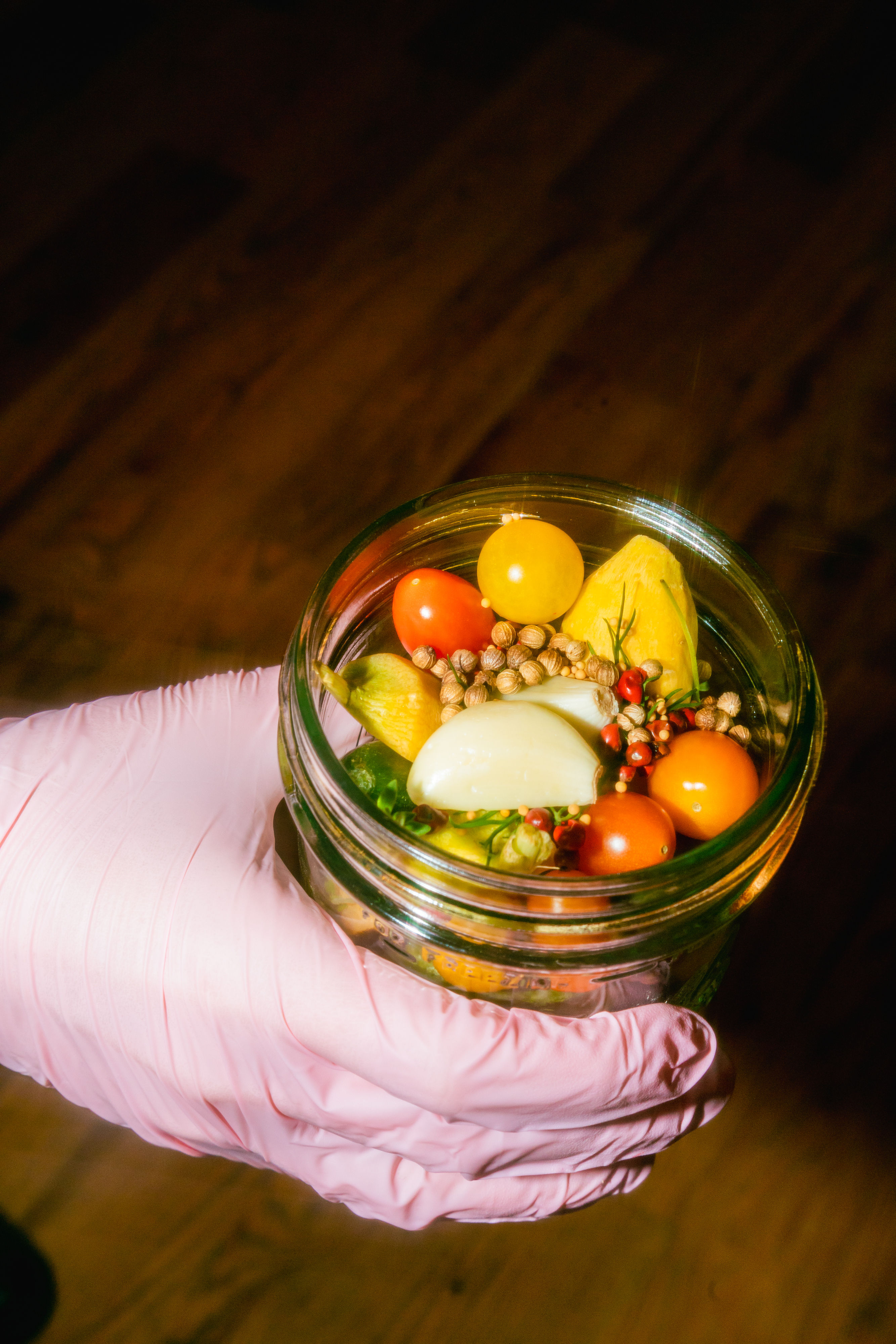 Close-up of a mason jar with vibrant pickled vegetables — yellow and red cherry tomatoes, a garlic clove, spices and herbs — held by a pink-gloved hand over a wooden surface.