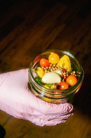 Close-up of a mason jar with vibrant pickled vegetables — yellow and red cherry tomatoes, a garlic clove, spices and herbs — held by a pink-gloved hand over a wooden surface.