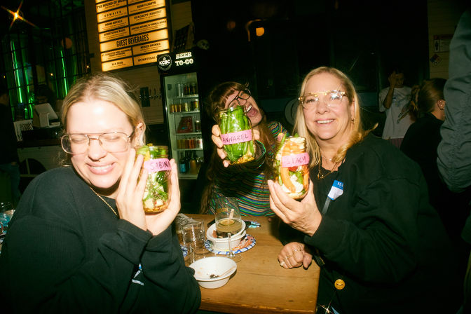 Three friends at a lively bar holding mason jars of layered salads and pickled veggies with name tags, wooden table with drinks, neon lights and beer fridge in the background
