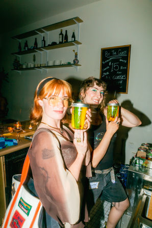 Two smiling people holding mason-jar cocktails at a cozy indoor bar counter, bottle-lined shelves and a chalkboard "By the Glass" menu in the background.