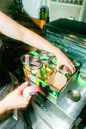 Hands labeling mason jars of pickled vegetables with colorful sticky notes on a stainless-steel prep counter next to a toaster oven in a busy kitchen.