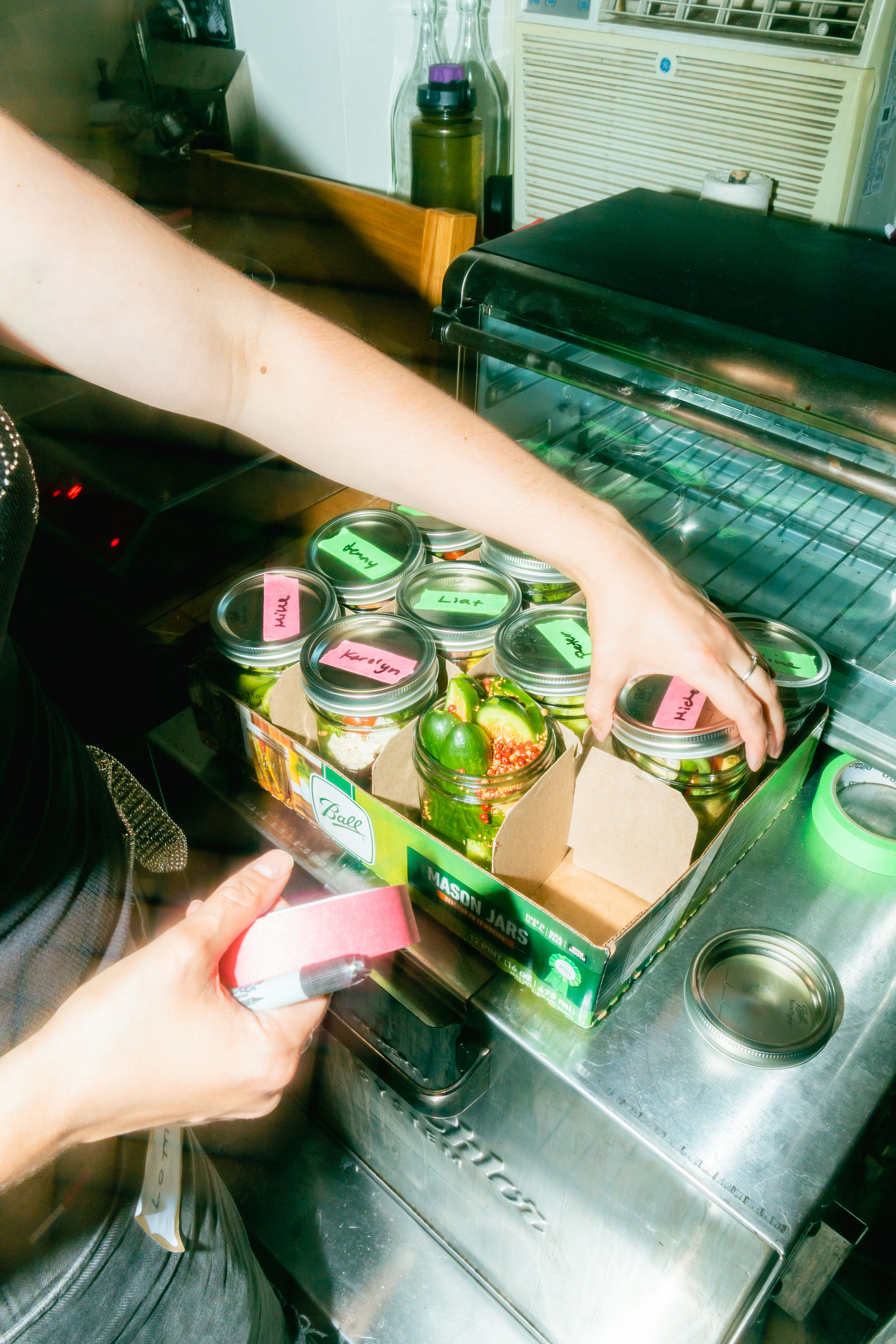 Hands labeling mason jars of pickled vegetables with colorful sticky notes on a stainless-steel prep counter next to a toaster oven in a busy kitchen.