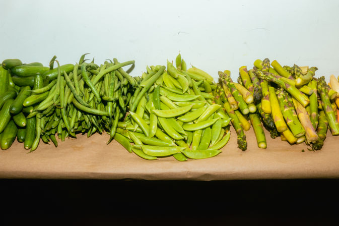 Fresh green produce on a market table — cucumbers, green beans, sugar snap peas and asparagus piled in bright rows.