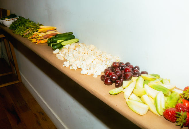 Colorful buffet-style spread of fresh fruit and vegetables on a long ledge: sliced apples and pears, cherries, strawberries, cauliflower, cucumbers and mixed carrots and snap peas — healthy party snack display