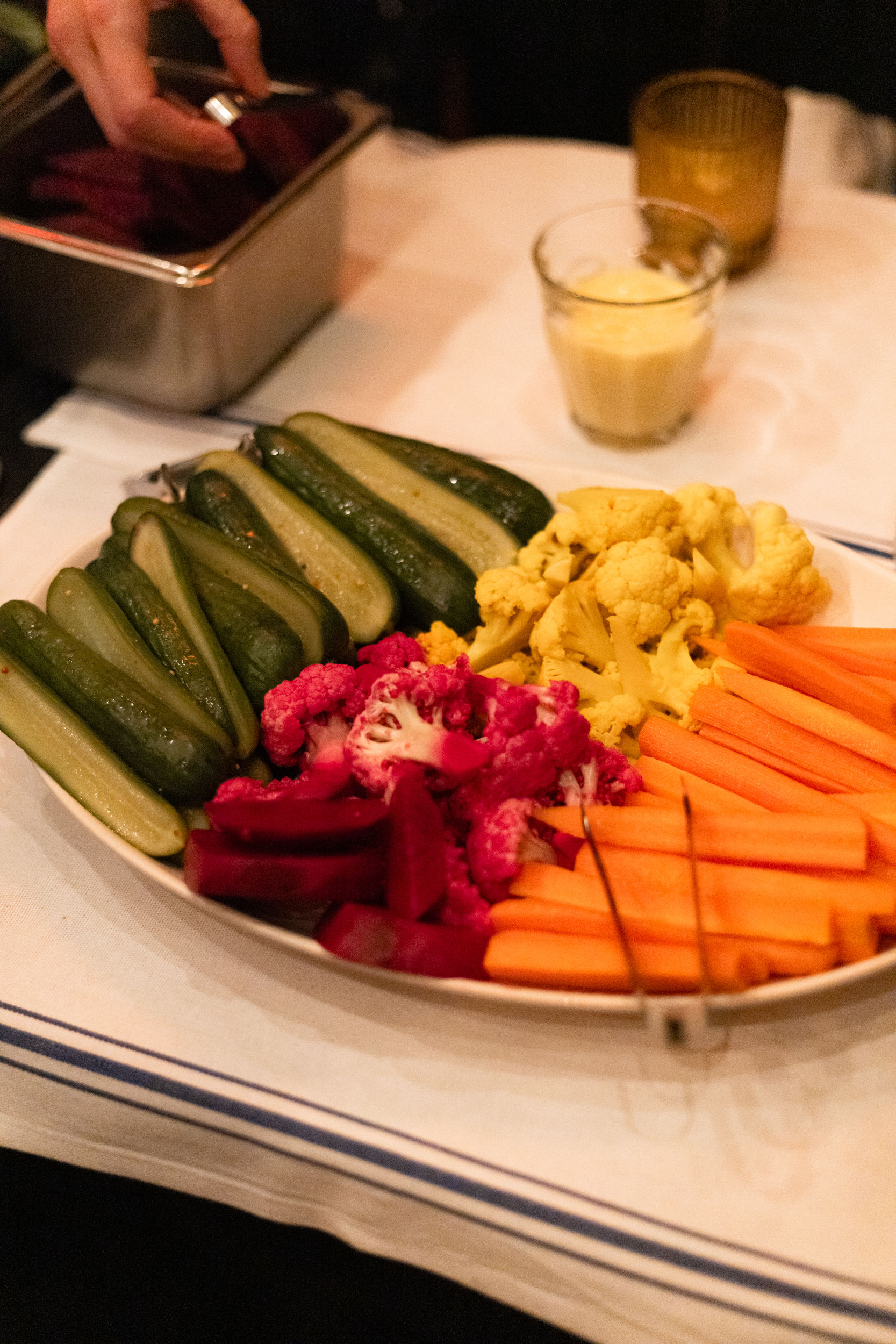 Colorful vegetable platter with pickles, pink and yellow pickled cauliflower, carrot sticks and beet slices next to a small glass of mustard dip on a white-tablecloth table, hand reaching with tongs.